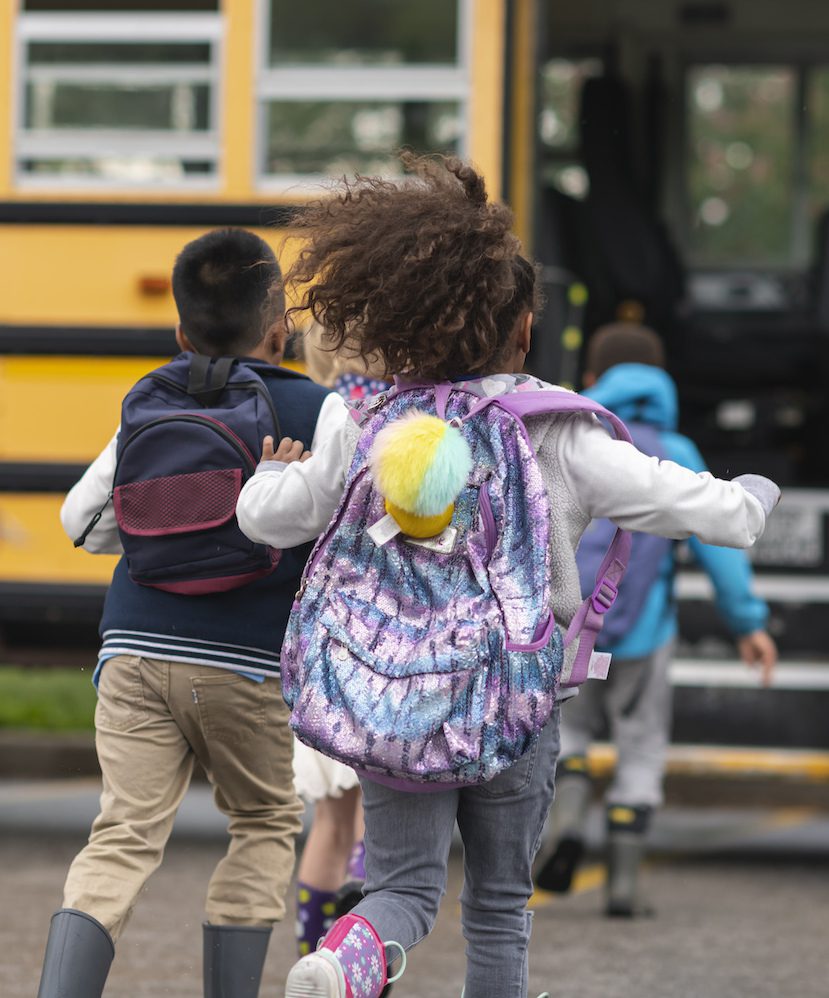 A multi-ethnic group of elementary age children are getting on a school bus. The kids' backs are to the camera. They are running towards the school bus which is parked with its door open. It's a rainy day and the kids are wearing jackets, rain boots and backpacks.